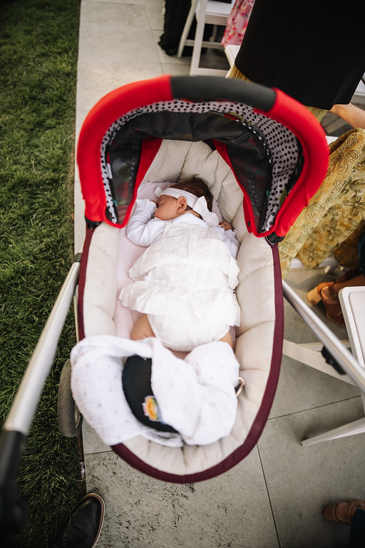 Overhead view of a sleeping baby lying in a stroller on a patio, surrounded by grass and guests’ chairs.