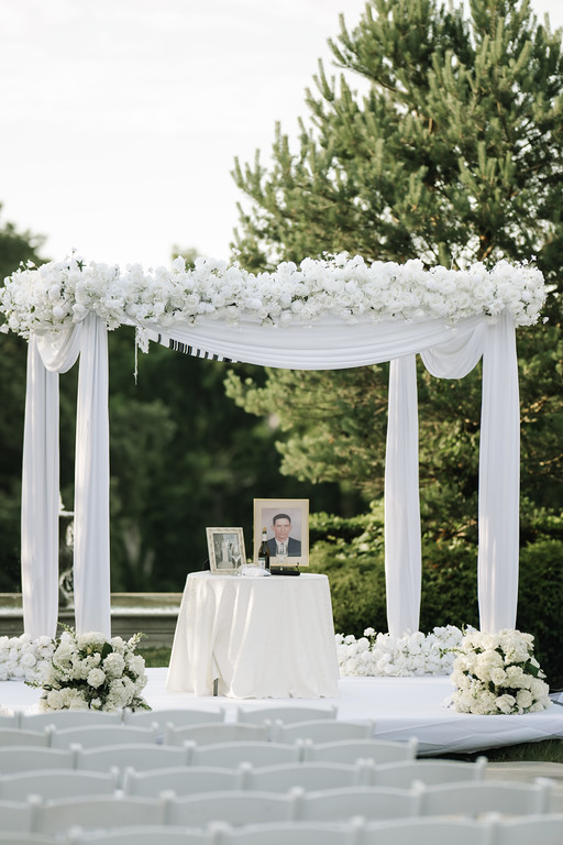 White floral chuppah with draped canopy and a table underneath featuring framed portraits at an outdoor wedding ceremony.