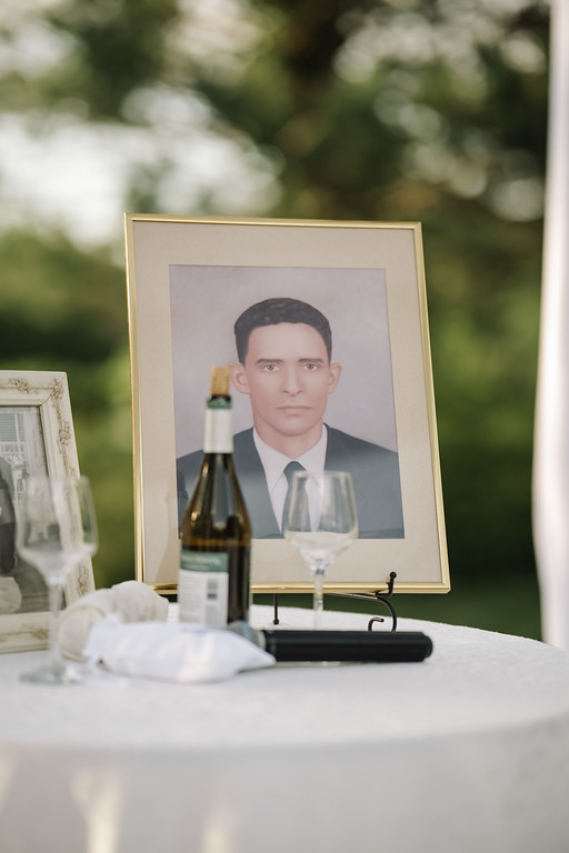 Framed portrait of a man placed on a table with wine bottle and glasses during a Jewish celebration, outdoors with green garden background.