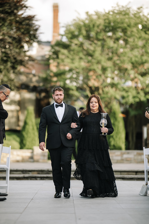 A couple in formalwear walking outdoors at a wedding event, smiling while the bride holds a glass in front of a garden backdrop.