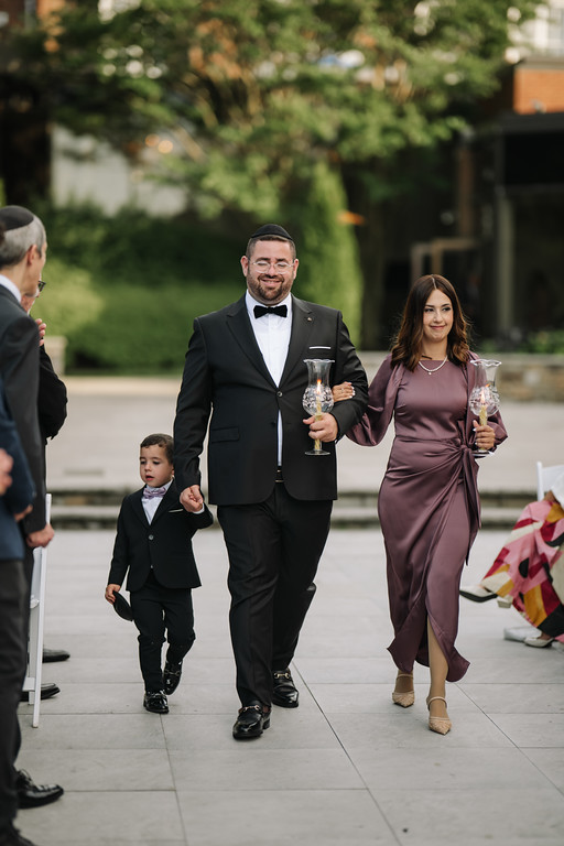 A groom in a black suit and kippah and a woman in a mauve dress walk side by side outdoors holding ornate ceremonial cups, with a toddler in formal clothing nearby.
