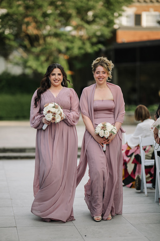 Two women in mauve dresses walk forward smiling while holding white bouquets at an outdoor wedding ceremony, with guests seated in the background.
