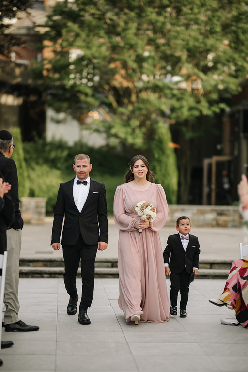 Bride in a blush gown holding a bouquet walks down an outdoor ceremony aisle with the groom in a black tuxedo and a young boy nearby.