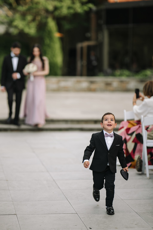 A young boy in a black suit and bow tie walks toward the camera smiling at a wedding venue, with seated guests and the couple in the background.