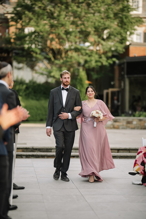 Bride in a blush-pink gown and shawl walking beside the groom in a dark tuxedo outdoors, holding a bouquet as a guest watches from the side.