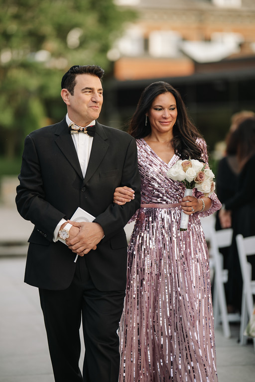 Framed couple outdoors at a wedding: groom in a black tuxedo and bow tie standing beside the bride in a sparkly pink gown holding a bouquet of white and pink roses.