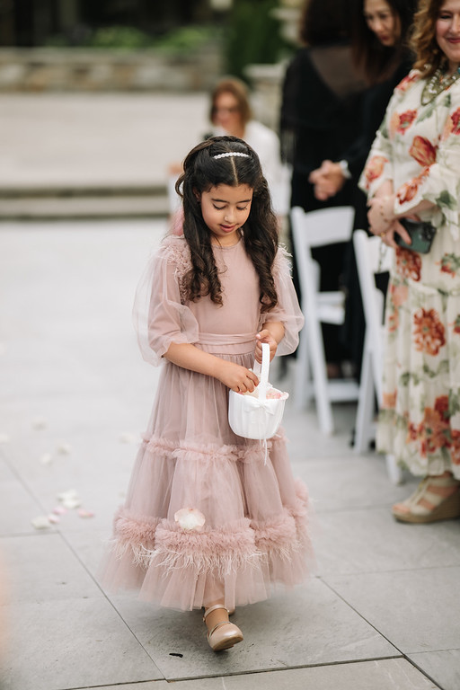 Young girl in a light pink dress and veil headpiece walking down the aisle while looking down at a small bag she is holding during a Jewish celebration.