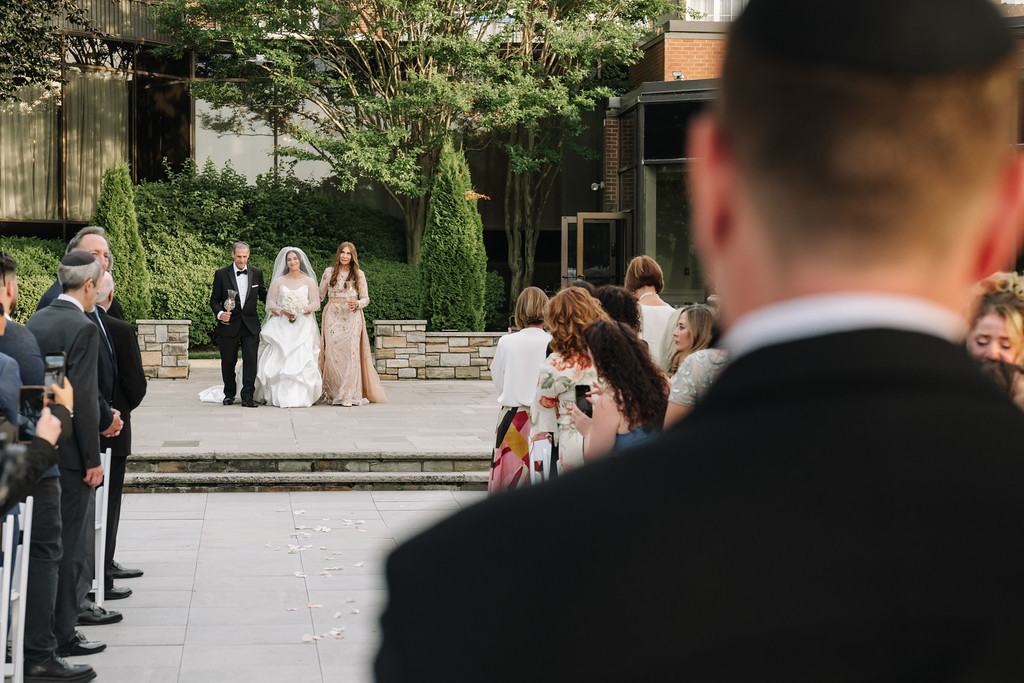 Outdoor courtyard wedding scene with the bride and groom standing on the steps near hedges, flanked by attendants as guests gather along the sides.