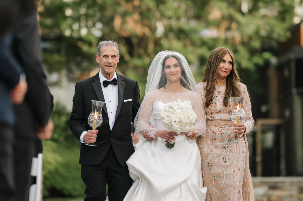 A bride in a white gown and veil holds a bouquet as she walks between her father in a tuxedo and her mother in an embellished dress outdoors during a wedding.