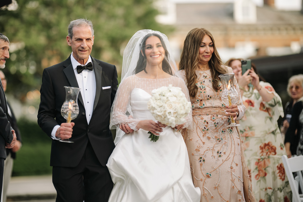 A smiling bride in a white gown and veil walks with her father and a woman holding her bouquet outdoors during a wedding procession.