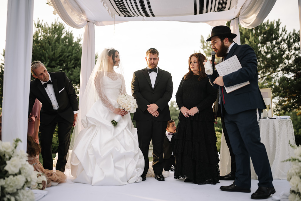 Wedding couple and family stand beneath a chuppah canopy as a rabbi reads from a siddur during the badeken/chuppah moment.