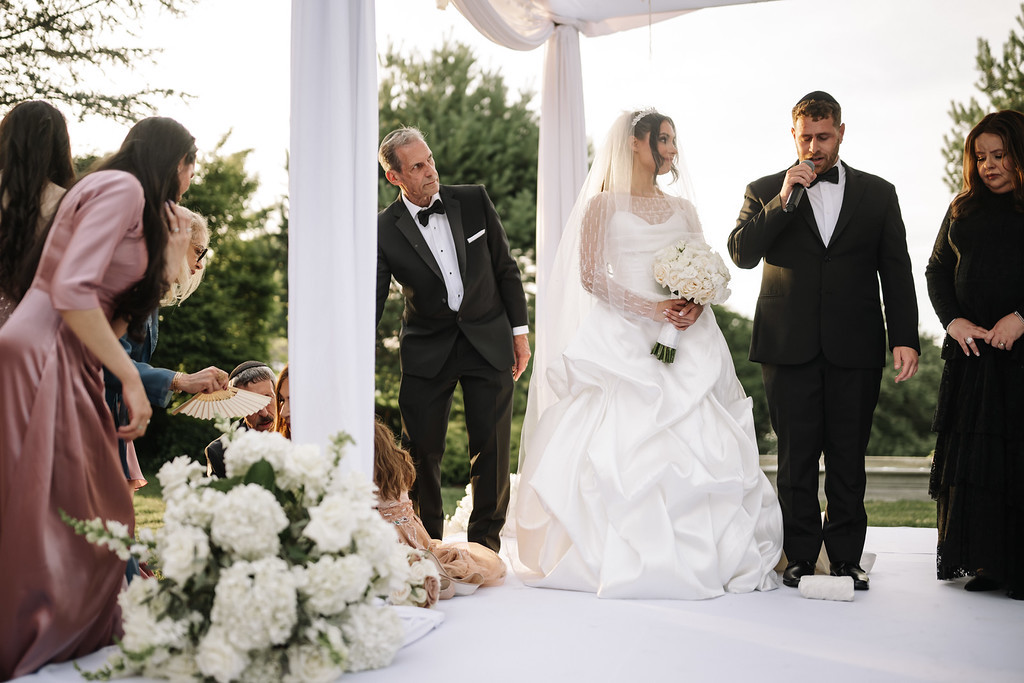Bride in a white gown under a chuppah canopy holding a bouquet, with the groom in a tux speaking into a microphone and family gathered on the aisle at sunset.