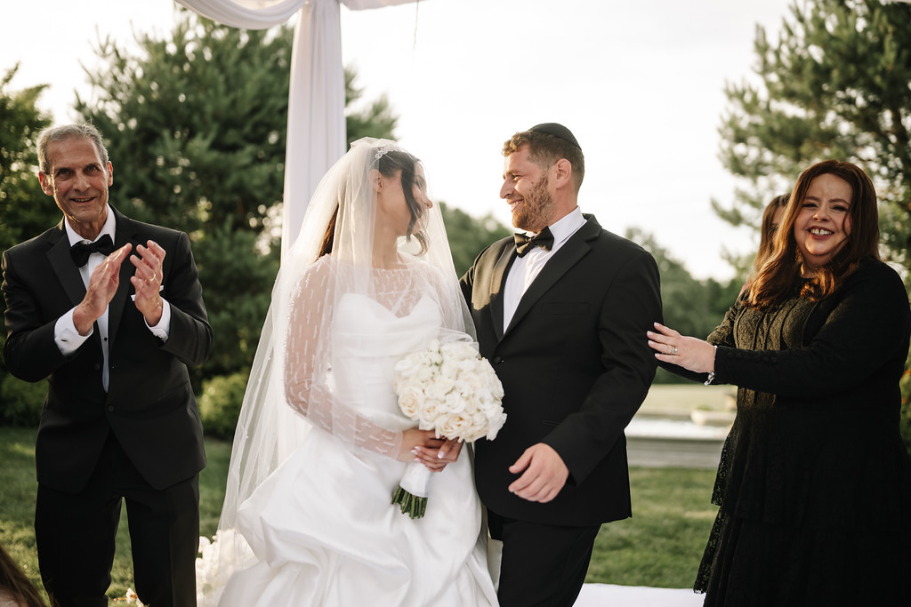 Bride in a white veil and dress holding a bouquet with the groom smiling beside her as guests clap outdoors under a white canopy.