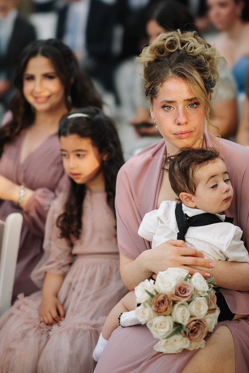 A woman in a soft pink dress holding a newborn in her arms, with a bouquet of pale roses in the foreground and other guests seated softly in the background.