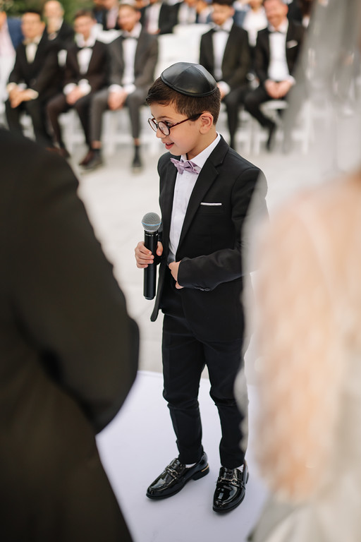 A young boy in a black suit and kippah holds a microphone and looks down while singing or speaking, with guests seated in formal attire behind him.