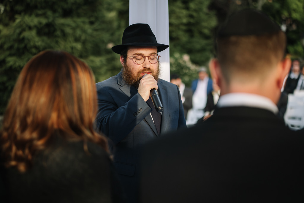 A man in a black hat and suit sings into a microphone at a wedding ceremony, with audience members in the foreground and greenery in the background.