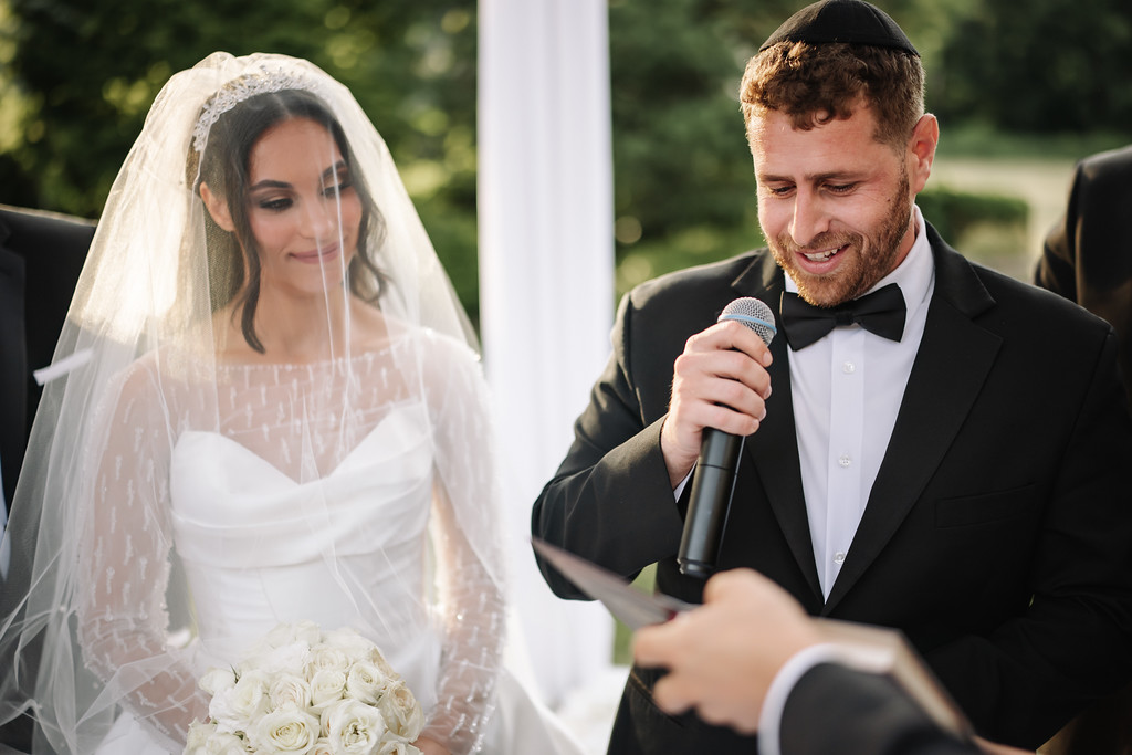 Bride in a white veil and lace gown stands beside the groom in a black tuxedo and kippah during the badeken, with the groom smiling while speaking into a microphone.