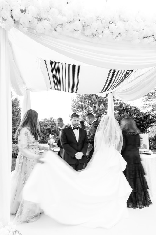 Black-and-white wedding moment under a canopy as the bride and groom stand together while guests and family move around in the background.