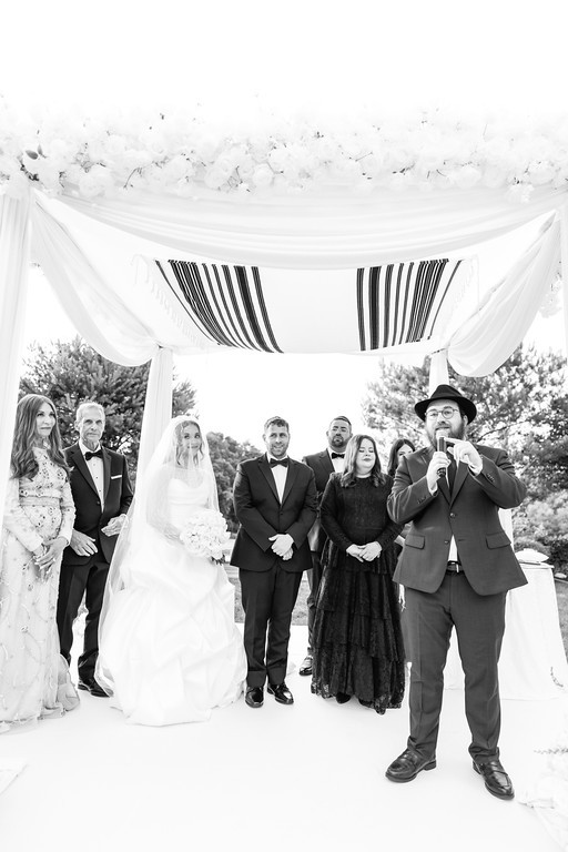 Black-and-white wedding scene under a chuppah with the bride and groom standing at center while family and guests line the walkway and a man in the foreground holds a camera and gestures.