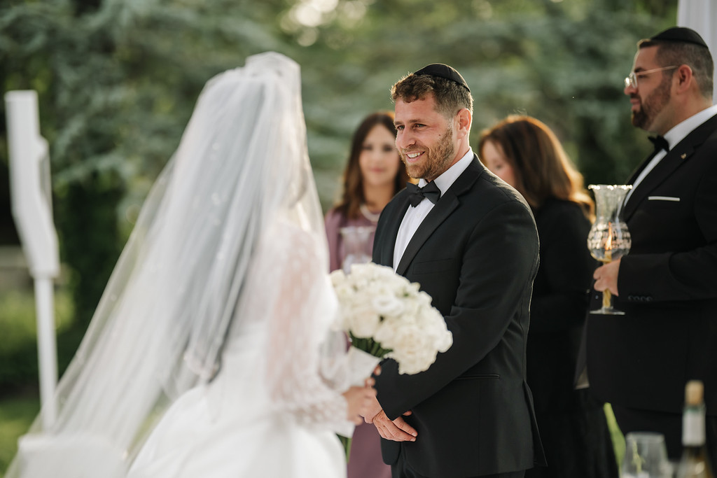 Groom in a black tuxedo smiling softly during badeken, holding the bride’s hands as the bride in a white veil faces him outdoors.