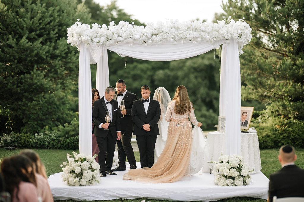 At an outdoor chuppah ceremony, the bride in a flowing gown walks into position under a white floral canopy as the groom and attendants stand together and guests watch.