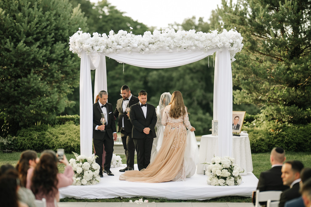 Bride and groom under a white floral chuppah during a Jewish wedding ceremony, with family and guests watching from the sides.
