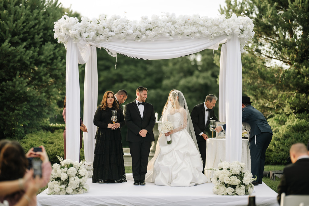 Bride and groom standing together under a white floral chuppah during an outdoor wedding ceremony, with attendants and family nearby.