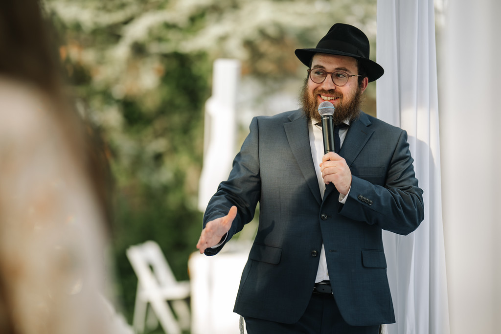 Smiling Orthodox Jewish man wearing a black hat and glasses speaks into a microphone under a white canopy, with greenery blurred in the background.