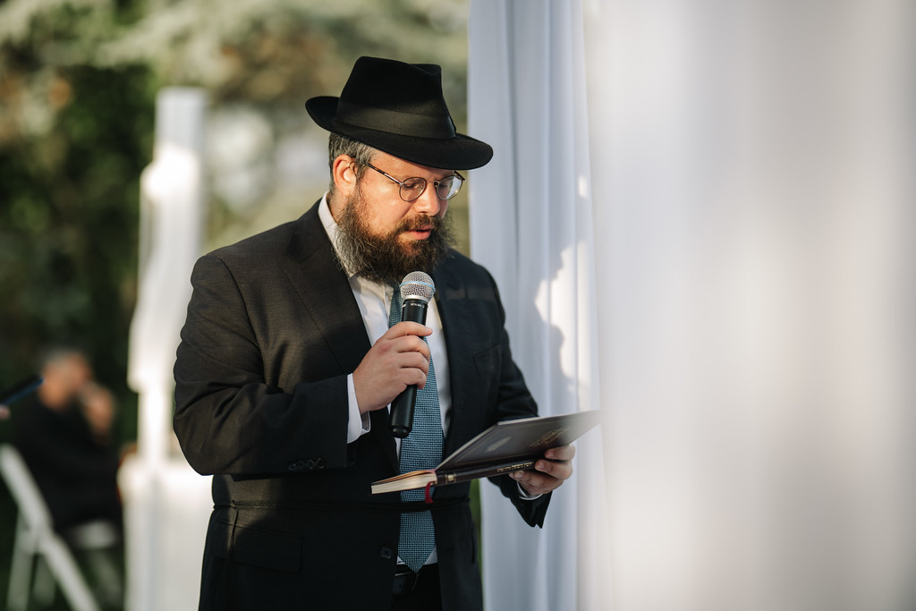 A bearded man in a black hat and glasses speaks into a microphone while reading from notes beside a white column at a Jewish event.