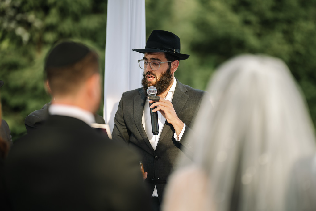 A bearded groom in a black hat and suit speaks into a handheld microphone at the chuppah, with the crowd softly blurred in the foreground and greenery in the background.
