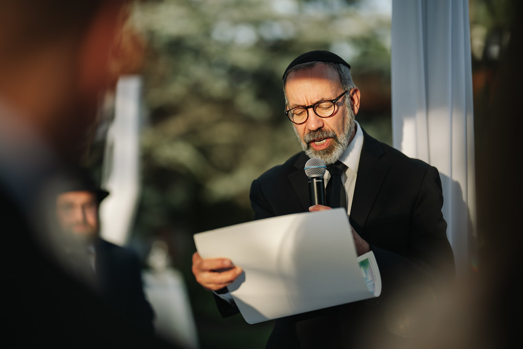 A bearded man wearing a black kippah and glasses reads from papers into a microphone at a wedding or event, with other guests softly blurred in the background.