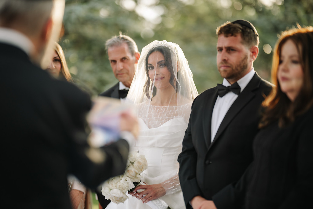 The bride in a white veil and dress at the badeken, standing among family and guests outdoors as the ceremony moment approaches.