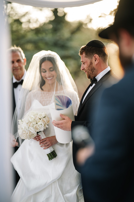 Bride in a white veil smiles while holding a bouquet and reading a program; groom in a black suit and kippah looks at her as guests watch in the background.