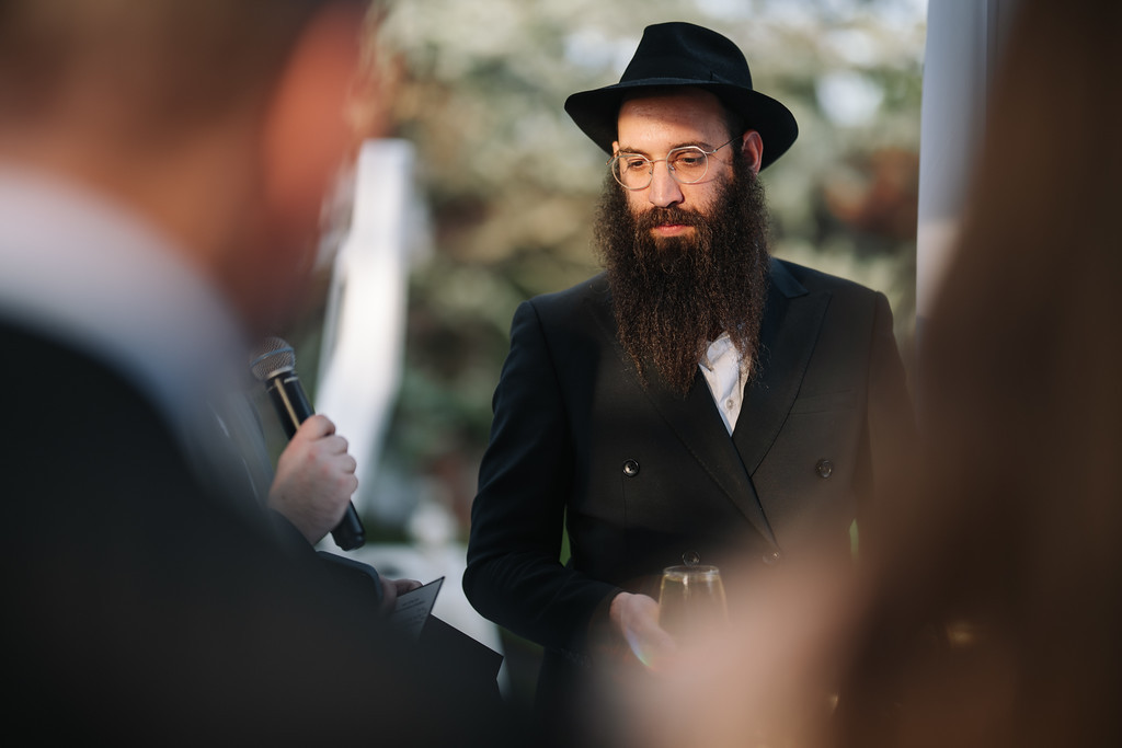 A man with a long beard and black hat wearing glasses stands outdoors in a suit during an event, speaking with a microphone while blurred people frame the foreground.