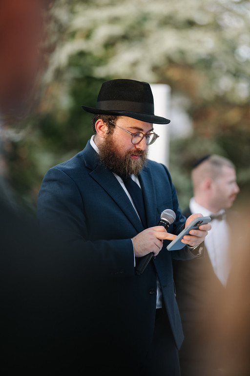 A bearded man in a black hat and suit speaks into a microphone while looking at his phone at an outdoor wedding celebration.