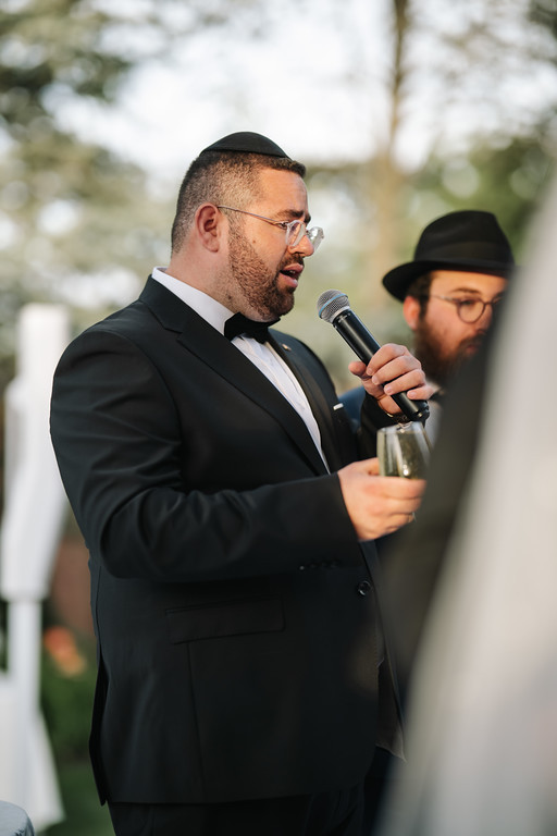 Frum wedding/ceremony speaker in a black suit holding a microphone, with another guest wearing a black hat and kippah watching in the foreground.