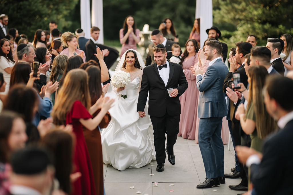 A bride in a white dress and veil and a groom in a black tuxedo walk together down the aisle under chuppah-style decorations while guests clap and smile around them.