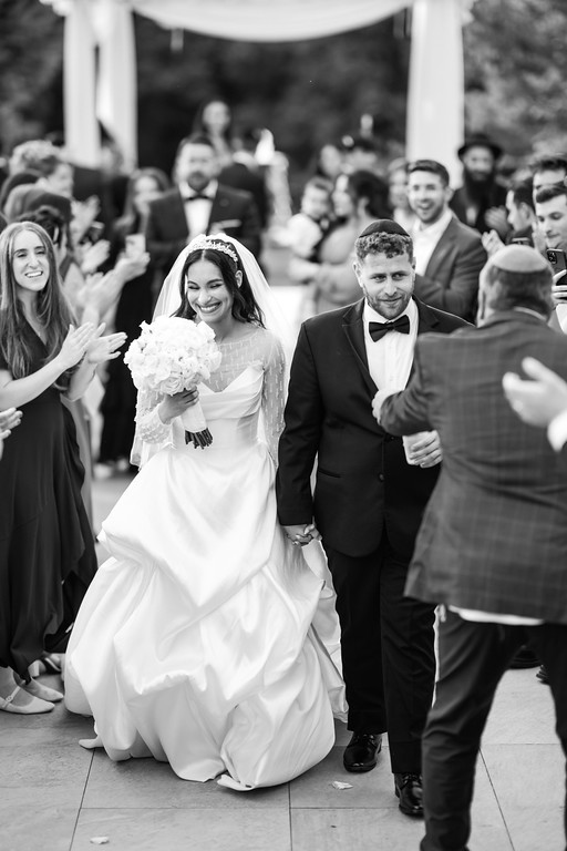Black-and-white wedding dance moment: the bride in a lace hood smiles while holding her husband’s hand as guests clap around the chuppah.