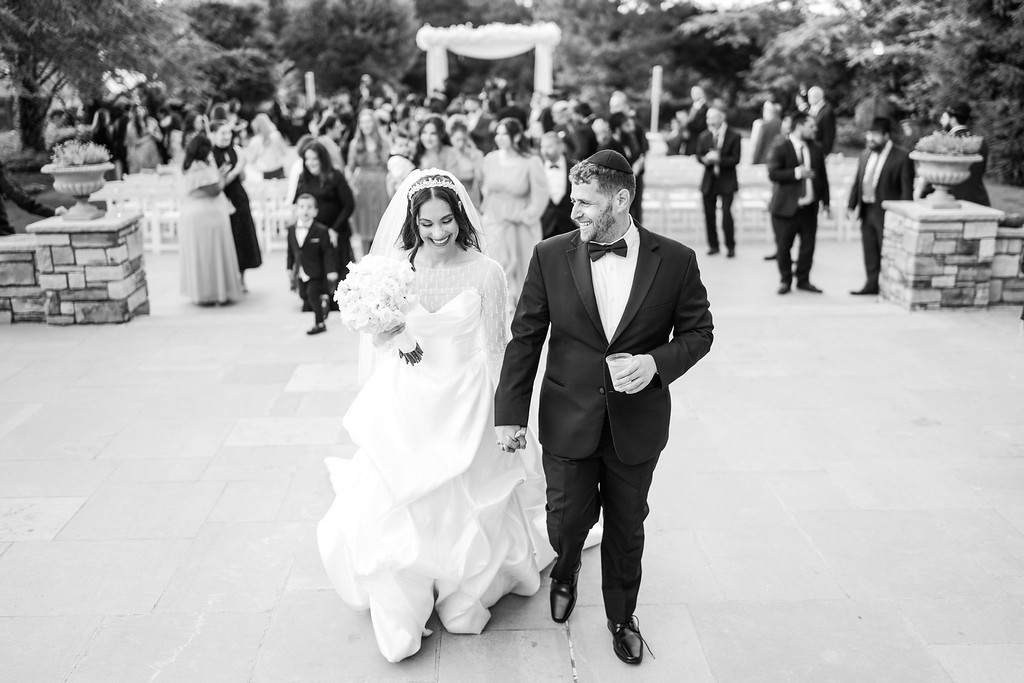 Black-and-white photo of a Jewish wedding couple walking together down an outdoor aisle after the chuppah, smiling and holding hands with guests and ceremony decor in the background.