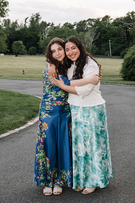 Two young women in colorful event dresses hug tightly and smile outdoors on a paved path with greenery in the background.