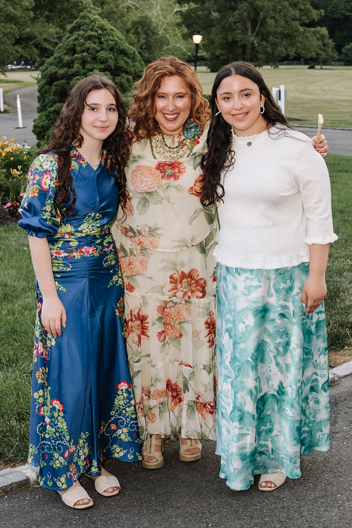 Three women stand side by side outdoors in a park, smiling—one in a floral dress, one in a blue patterned dress, and one in a light blue floral skirt and white top.