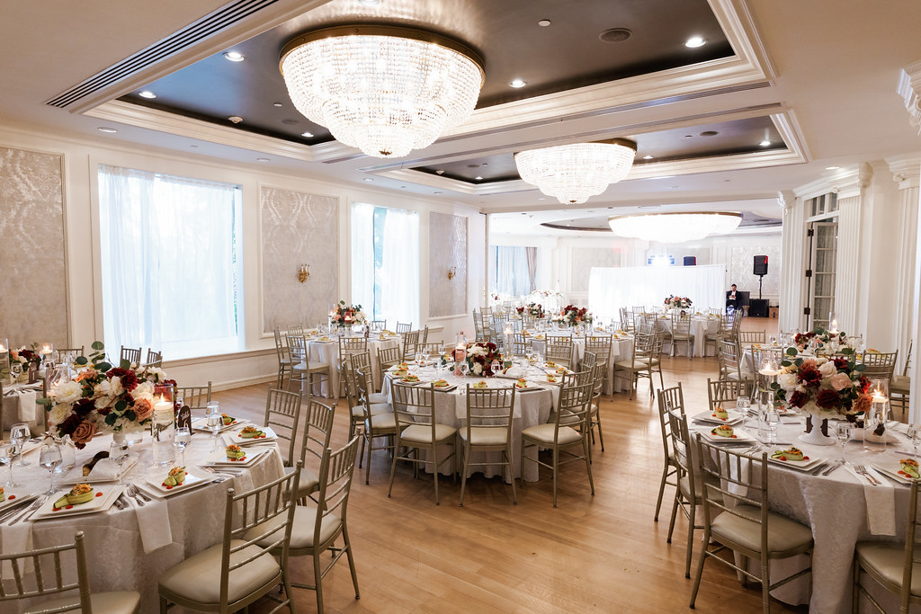 Reception hall interior with neatly set round tables, floral centerpieces, and chandeliers under bright windows.