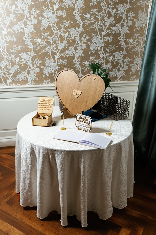 Close view of a wedding details table with heart-shaped decorations, small framed signs, and an open guestbook on a round cloth-covered surface.