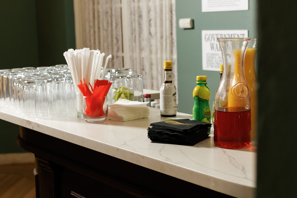 Close-up of a venue refreshment table with cups, napkins, bottled drinks, and a decanter against a wall.
