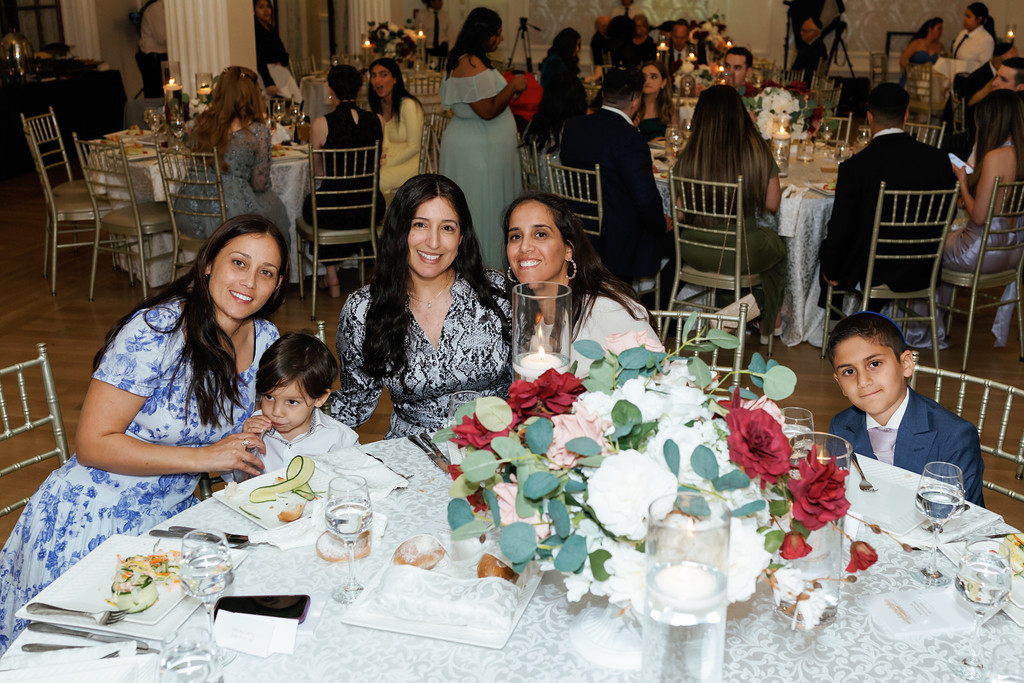 Two mothers and two children seated at a wedding reception table smile at the camera in front of a large floral centerpiece.