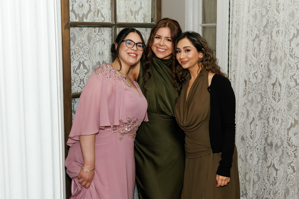 Three women standing close together in front of lace curtains, smiling at the camera—one in a pink dress, one in a dark green dress, and one in an olive top with dark outerwear.