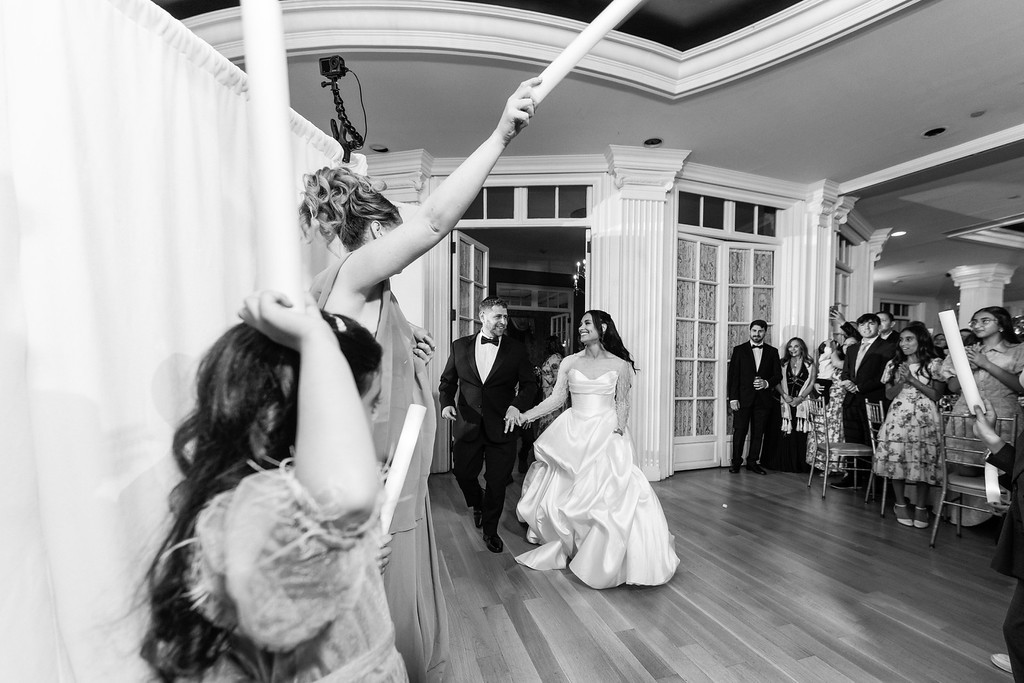 Black-and-white photo of a bride and groom entering a reception hall, smiling as guests on both sides reach toward them in celebration.
