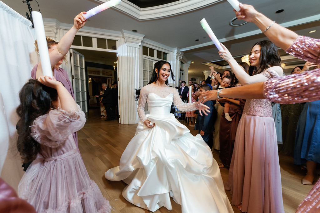 A bride smiles while dancing in a ballroom as guests in pastel dresses swing sparklers and cheer around her.