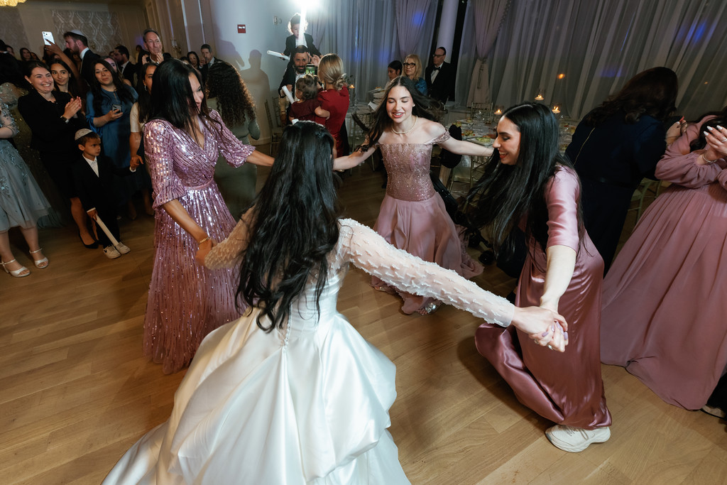 A group dancing at a wedding, with a smiling young girl in a sparkly dress holding her skirt while others stand around the dance circle in evening light.