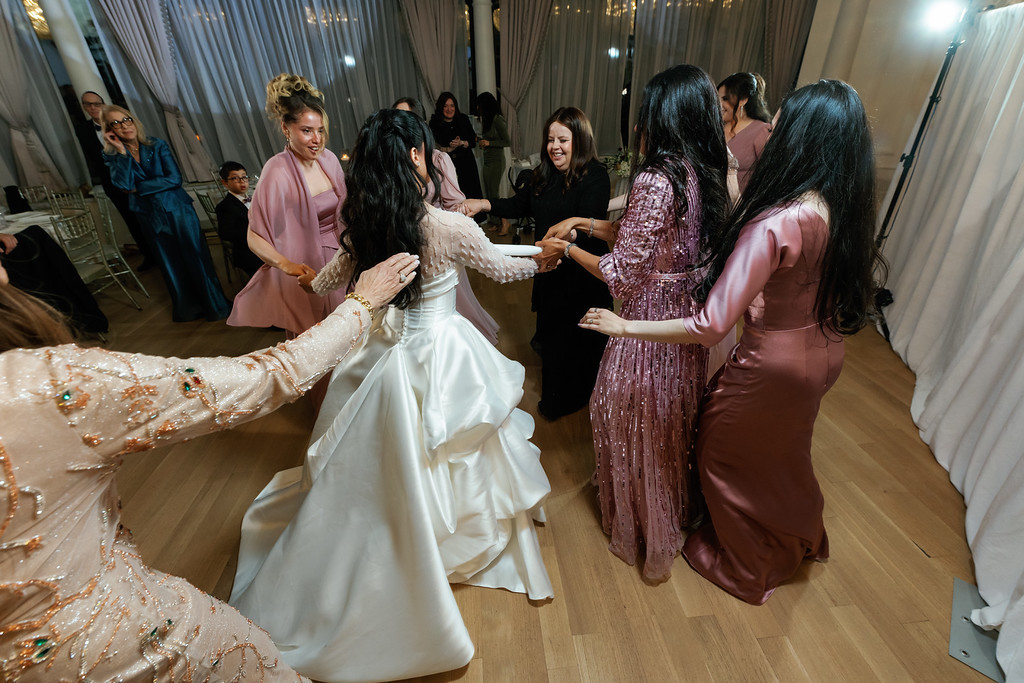 Bride in a white gown being lifted on the dance floor while guests surround her and smile at a wedding celebration.